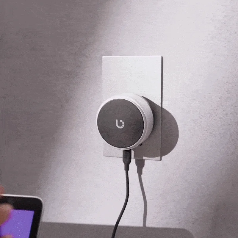 壁のコンセントにU字型ロゴのスマートデバイスが接続されています。充電器かスマートホームハブのような電子機器で、手前には画面の一部が見えます。