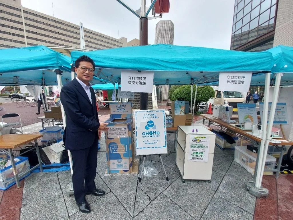 守口市役所主催の「OHOHO CYCLE PROJECT」の屋外イベント風景。スーツ姿の男性が小型家電の回収ボックスの横に立ち、環境保護とリサイクルを呼びかけている。参加者にはお菓子のプレゼントもある。
