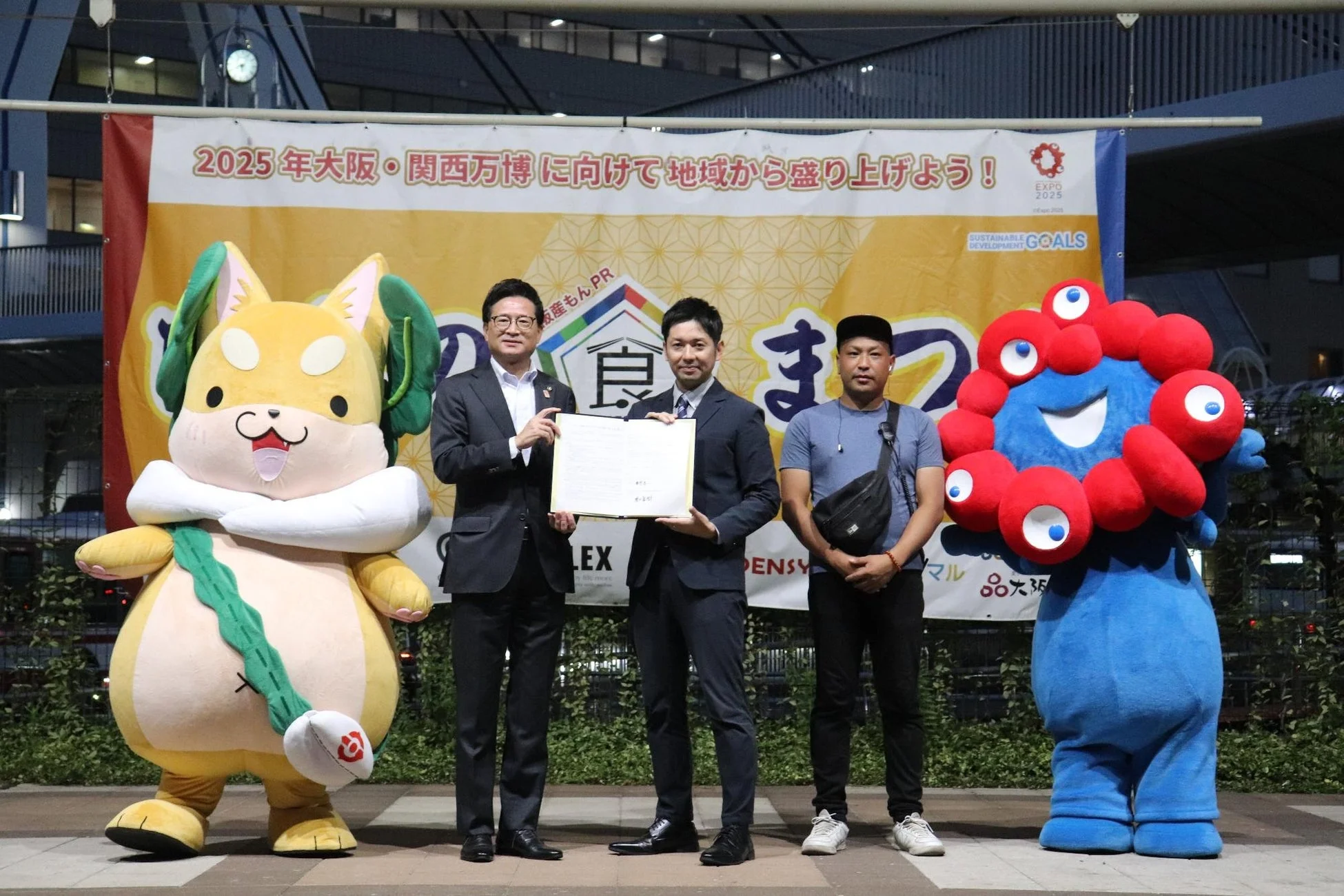 2025年大阪・関西万博の公式キャラクター「ミャクミャク」と「モッピー」を含む3人の男性が、万博を盛り上げるためのイベントで記念撮影している様子。背景には万博開催を呼びかけるバナーが見える。