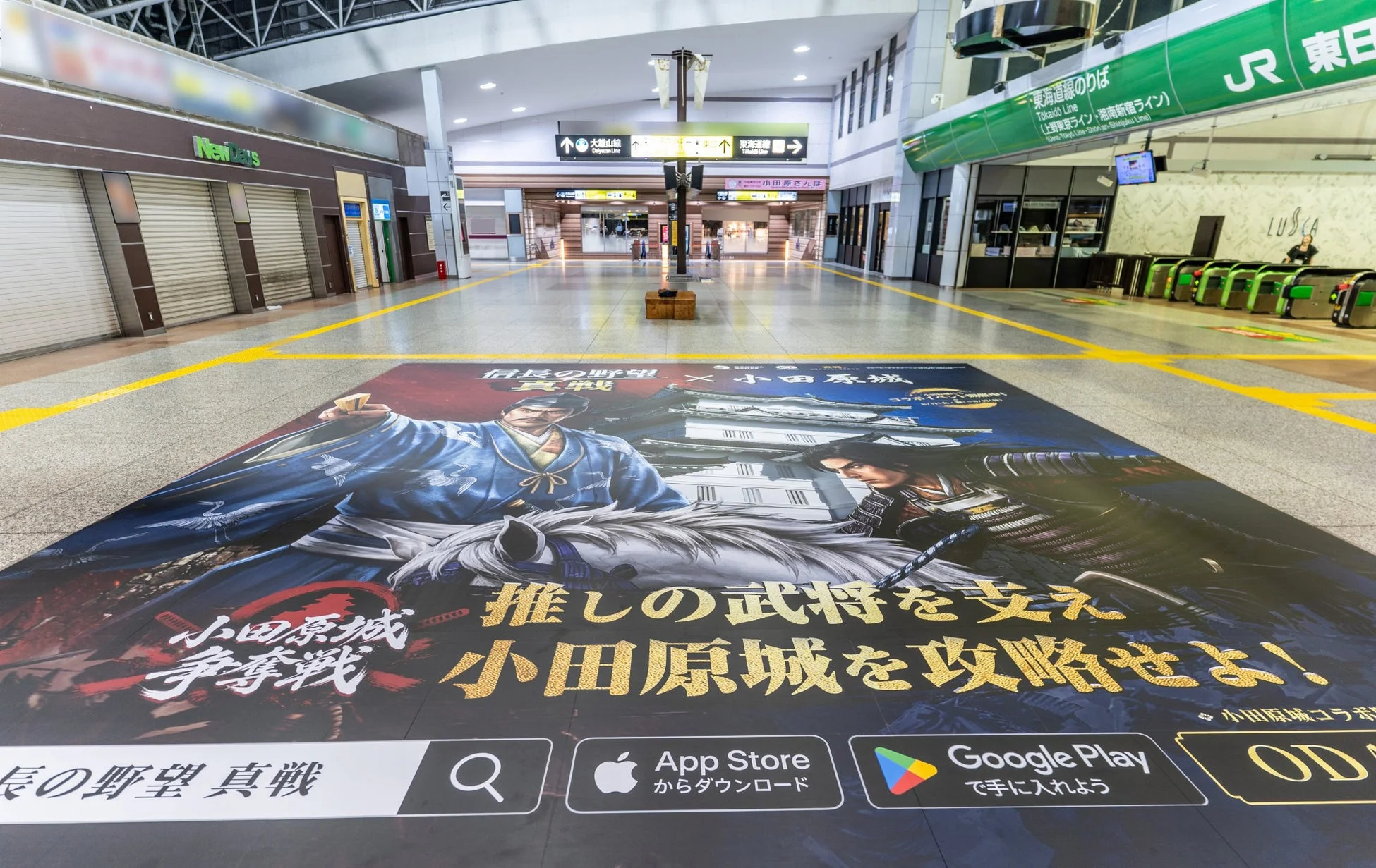 広々とした駅構内の床に、戦国ゲーム「信長の野望 真戦」と小田原城のコラボ広告が大きく掲示されている。武将のイラストと小田原城攻略を促すメッセージ、アプリストアへの誘導が特徴。JR改札やNewDays店舗も見える。