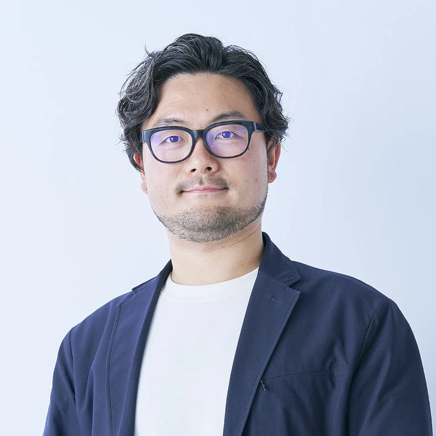 眼鏡とヒゲが特徴のアジア人男性のポートレートです。紺色のジャケットと白いTシャツを着用し、穏やかな表情でカメラを見ています。