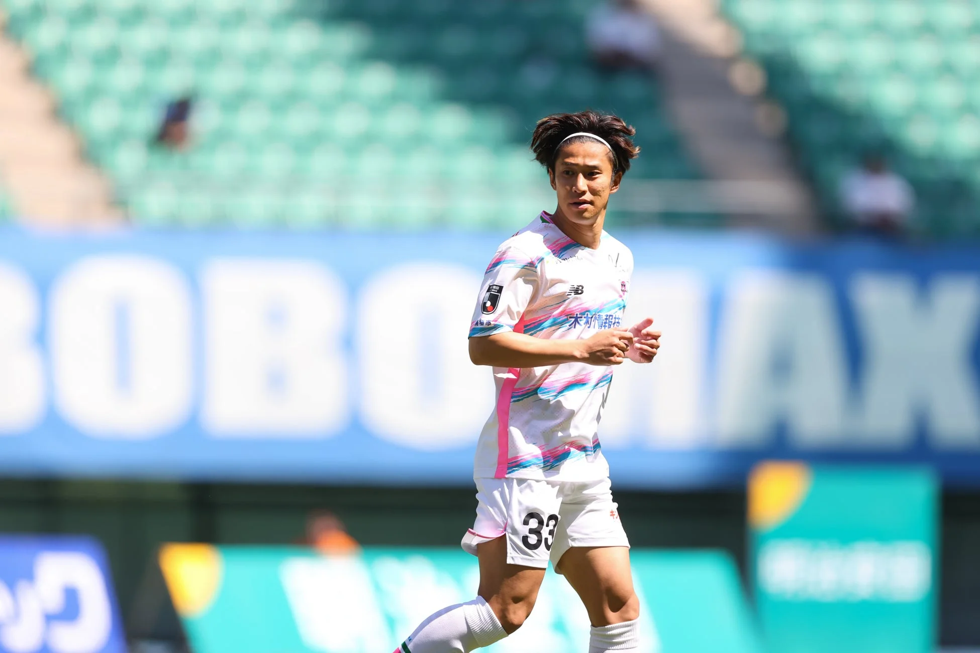 晴れたサッカー場で、背番号33の若い男性選手が白いユニフォームを着て走っています。ユニフォームにはピンクと水色のストライプ模様があり、試合中の真剣な様子がうかがえます。