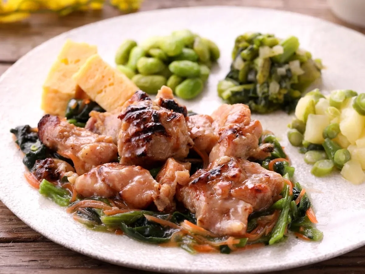 グリルされた鶏肉を主菜に、枝豆、青菜、ジャガイモとインゲンのサラダ、卵焼きのようなものが添えられた、彩り豊かな和風プレート料理