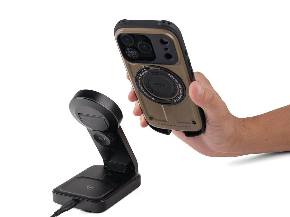 カーキ色の耐衝撃スマホケースを装着したスマートフォンを手に持ち、その手前には黒いワイヤレス充電スタンドが配置されています。MagSafe対応のモバイルアクセサリーを示す一枚です。