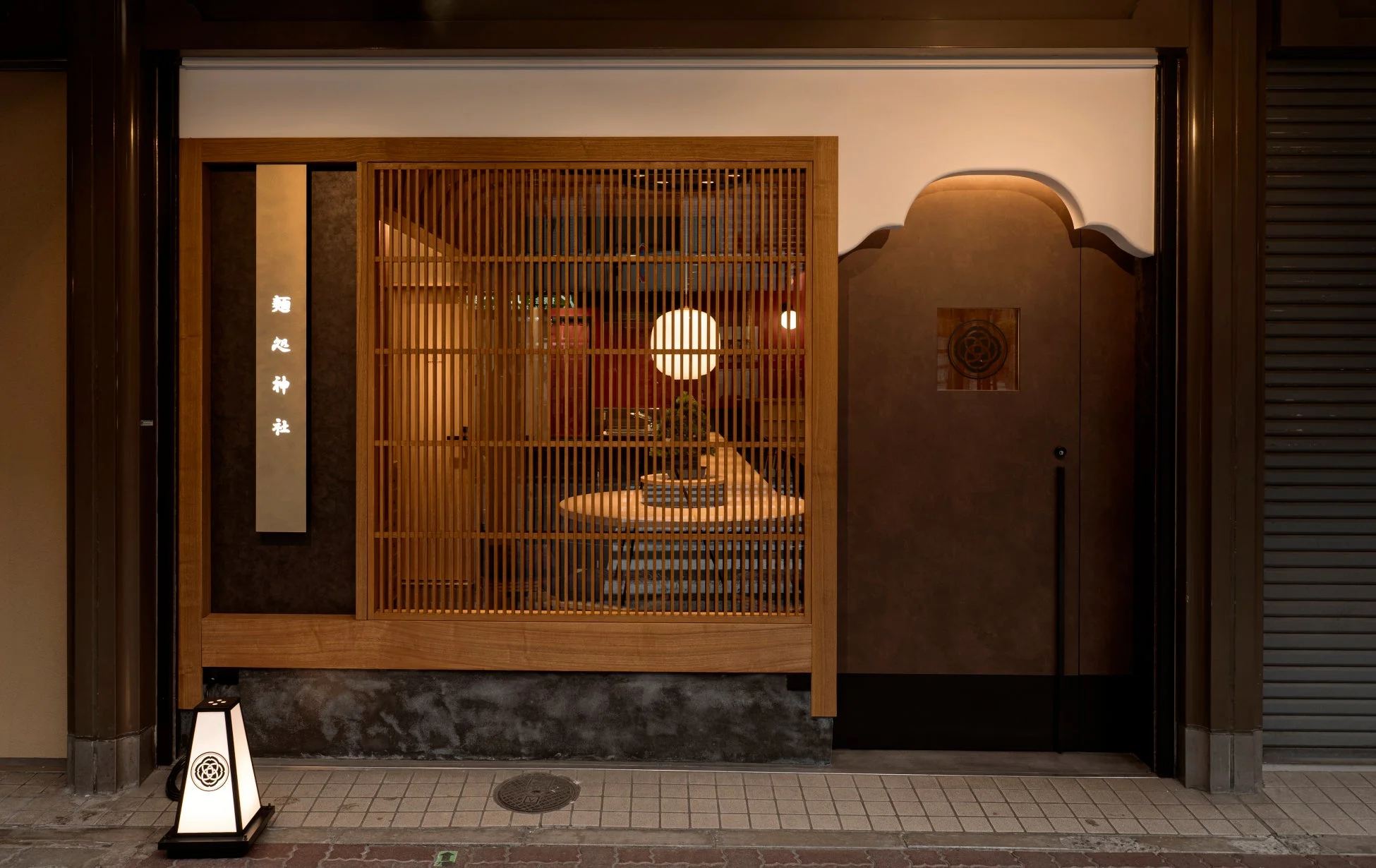 伝統的な和風建築とモダンなデザインが融合した店舗のファサード。木製の格子戸や提灯、店内の照明が温かい雰囲気を醸し出し、「麺処 神社」という文字が見えます。