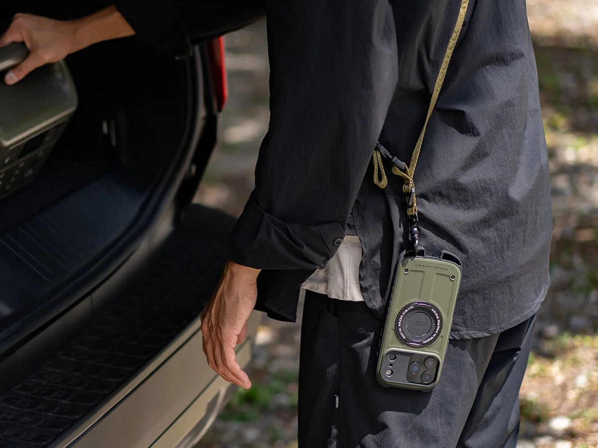 オリーブグリーンのタフネススマホケースを首から下げた人物が、車の荷台に物を積み込んでいる様子。SHOCK RESISTとROOT CO.のブランドロゴが見える。アウトドアシーンでの利便性を示す一枚。
