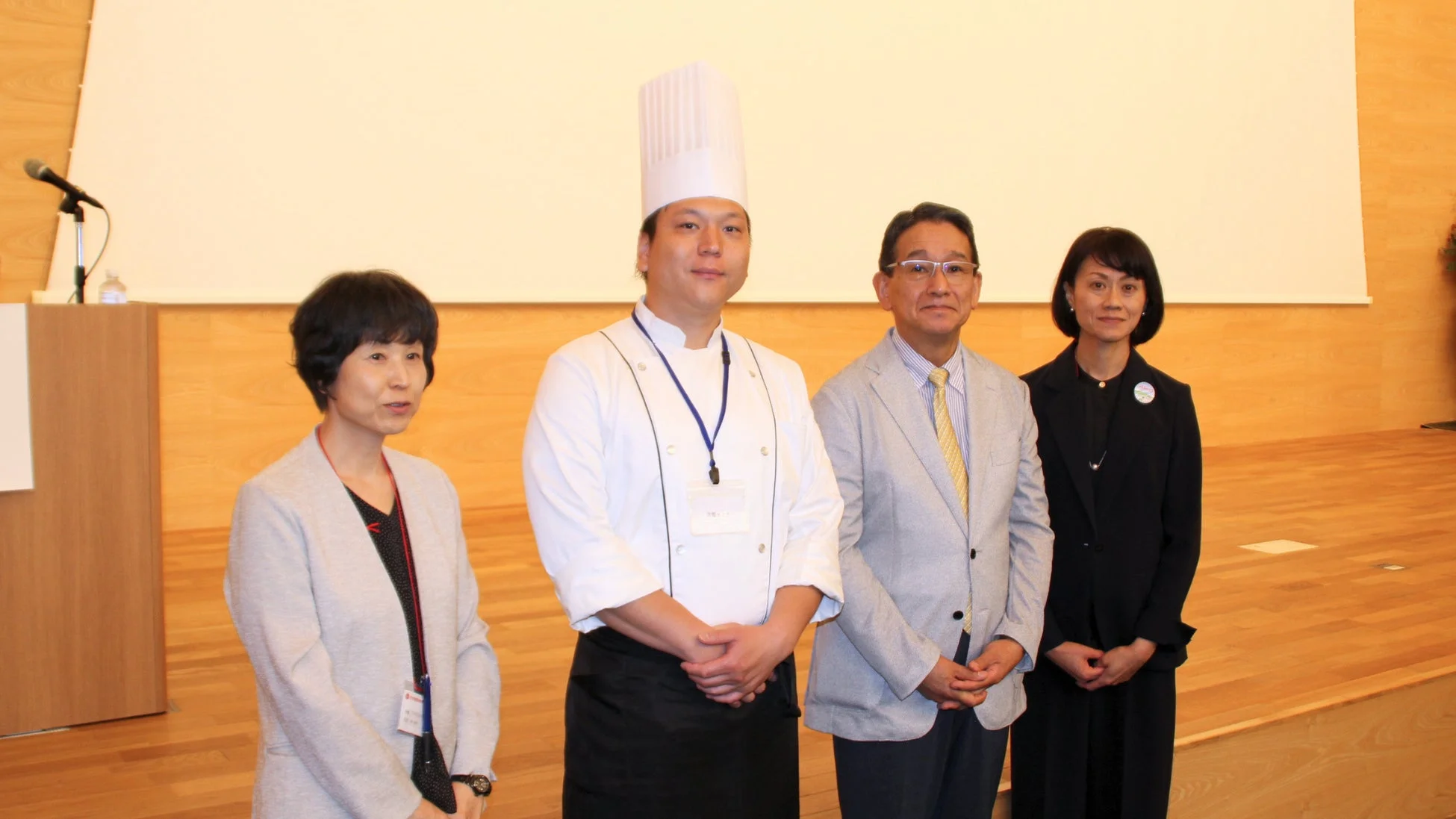 イベント会場で、シェフを含む4人の男女が並んで立っている集合写真です。背景には大きなスクリーンが見え、セミナーや講演会の一幕のようです。