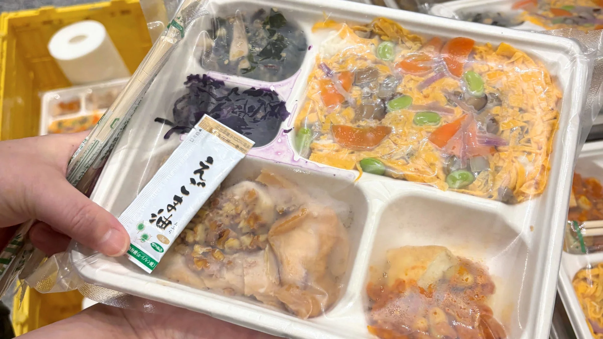 手元に持たれた冷凍弁当の画像です。複数の区画に分かれたおかずとご飯、えごま油の小袋、箸が確認できます。手軽に食べられる和風の健康的な食事であることが伺えます。