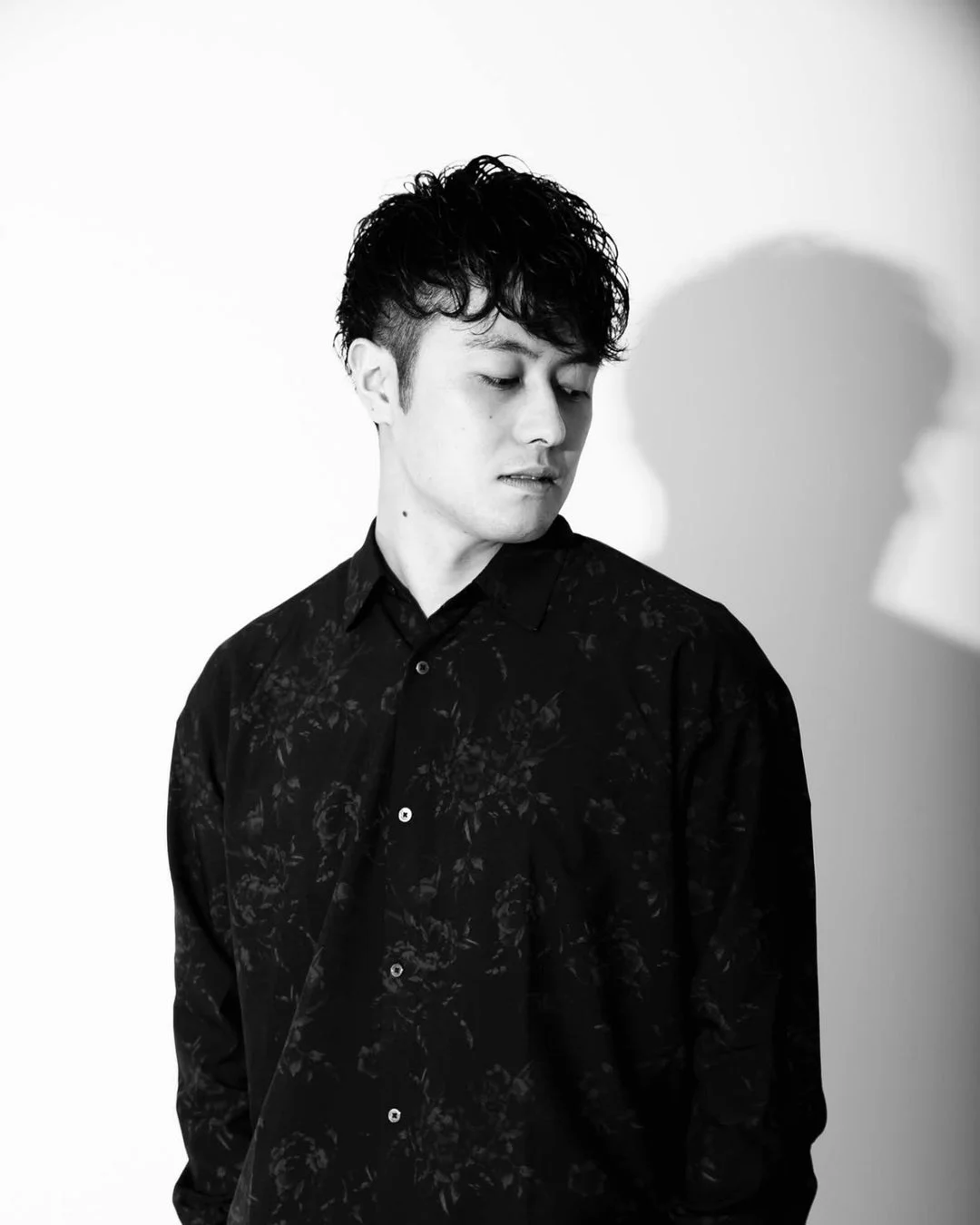 モノクロで撮影された若い男性のポートレート。彼は花柄の黒いシャツを着ており、目を伏せて思慮深い表情を見せている。