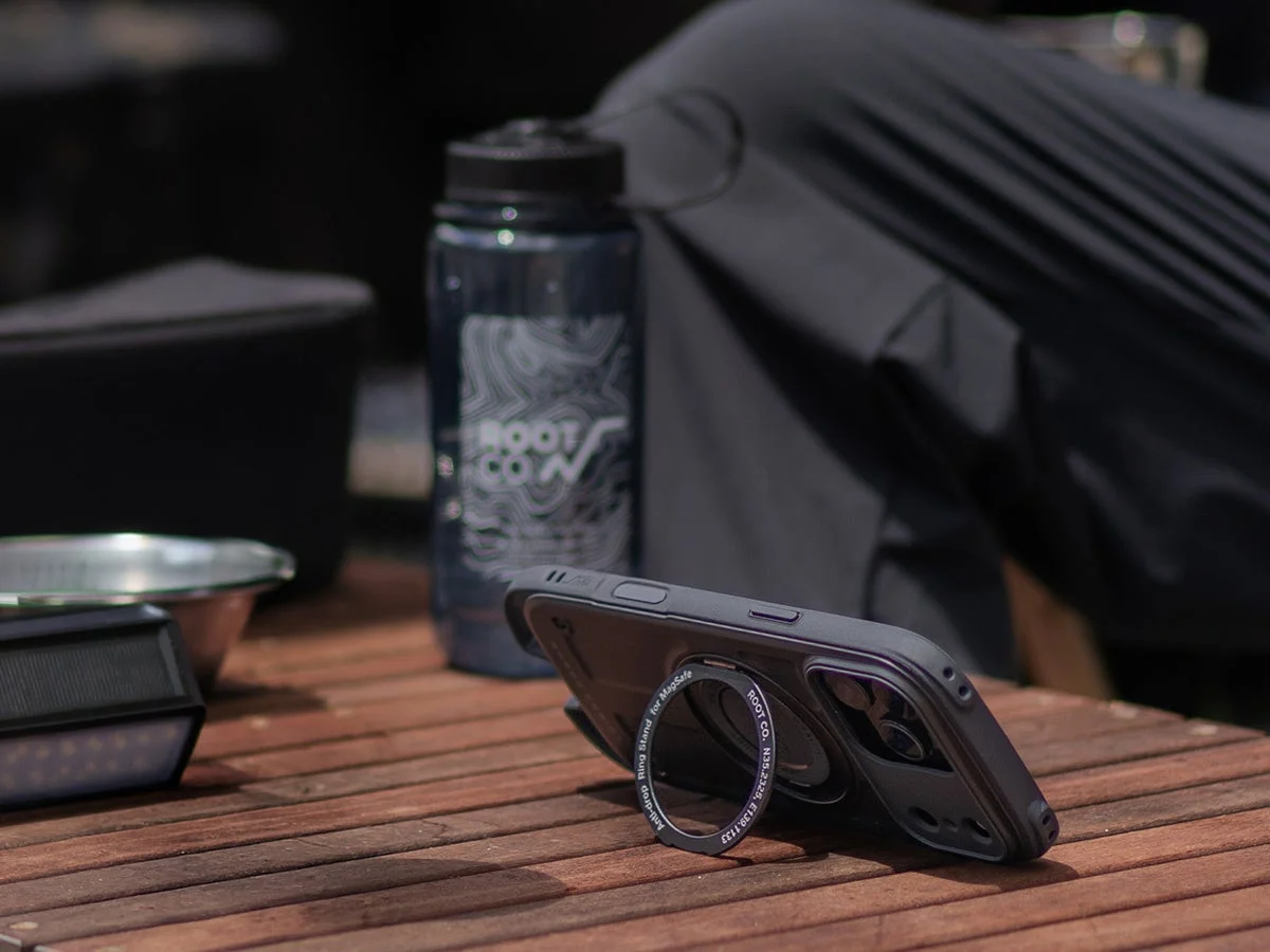 木製のテーブルに置かれたスマートフォンと水筒を捉えた画像です。スマホにはMagSafe対応のリングスタンドが装着されており、水筒とリングスタンドには「ROOT CO.」のロゴが見えます。アウトドアでの使用を想起させるシーンです。