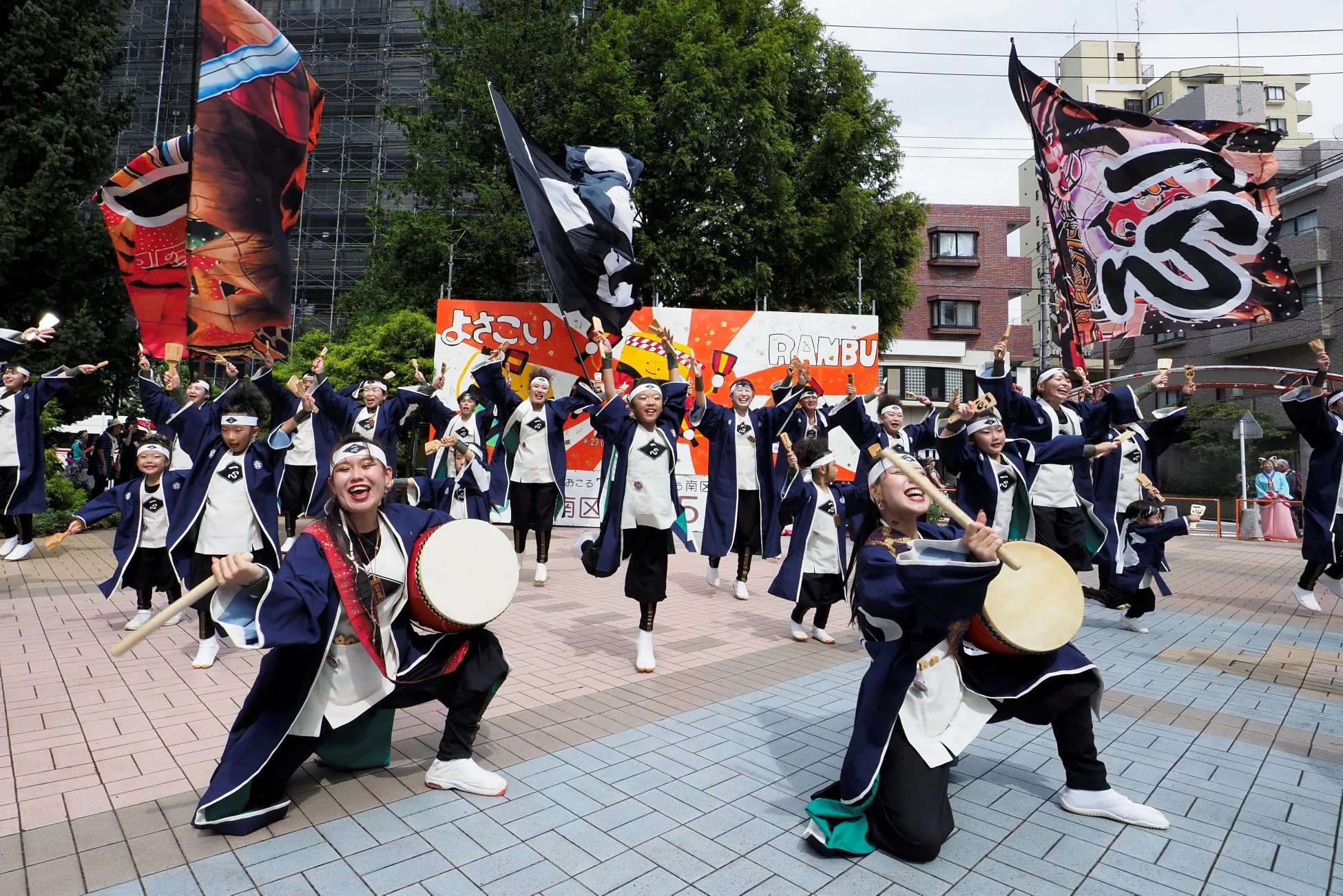 青と白の衣装を着た人々が、太鼓や鳴子を手に笑顔で踊るよさこい祭りの様子