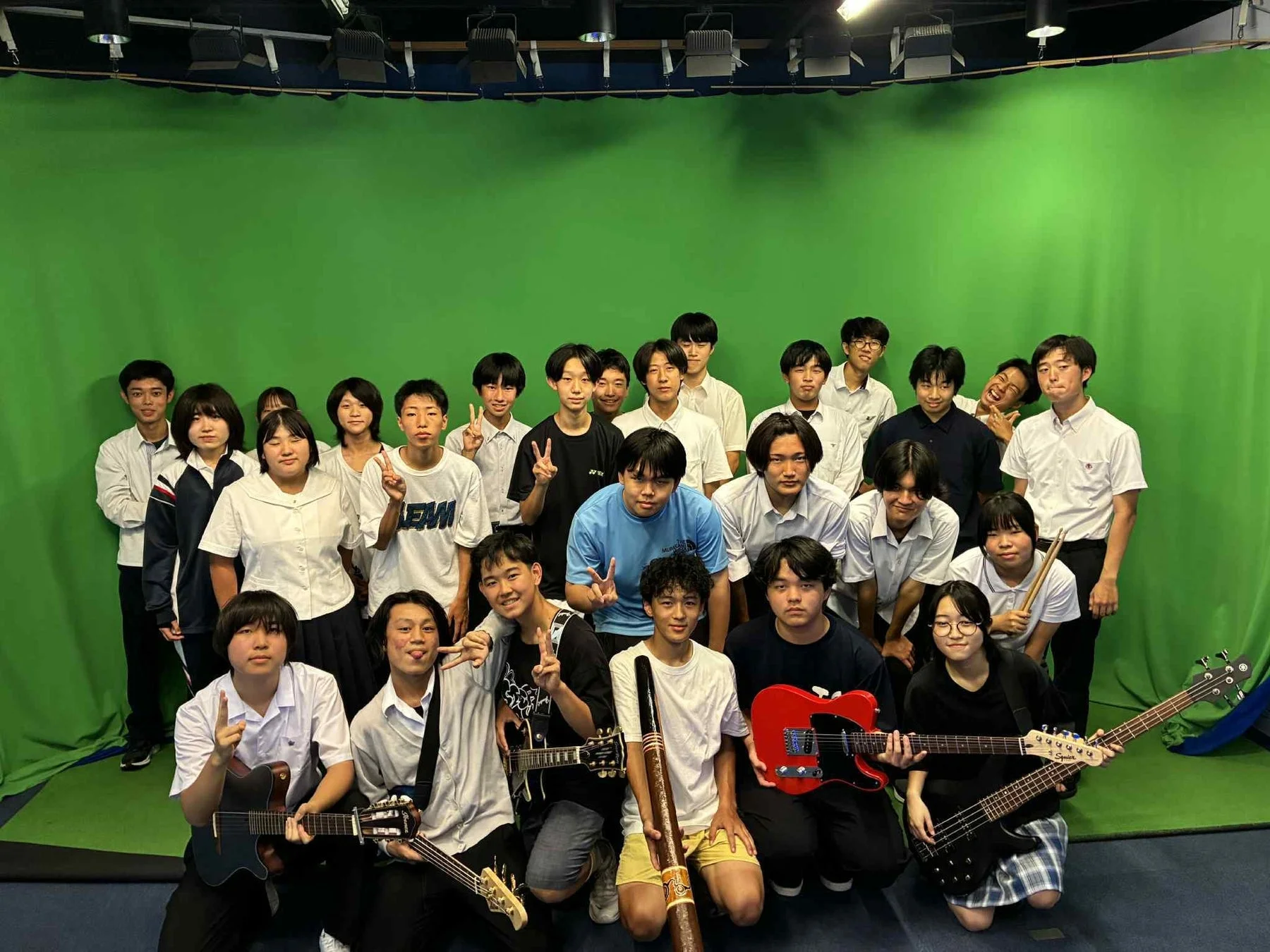 グリーンスクリーンを背景に、多数の学生がギターやベースなどの楽器を持って集合写真を撮っています。