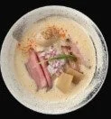 クリーミーなスープのラーメンが鉢に盛られています。薄切りの豚肉(チャーシュー)、刻んだ赤玉ねぎ、メンマがトッピングされ、赤い香辛料が振りかけられています。