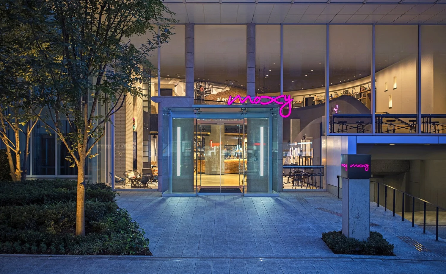夜にライトアップされたモダンな建物のエントランス。ピンク色の「moxy」ネオンサインが目を引き、ガラス越しにスタイリッシュなロビーが広がっています。