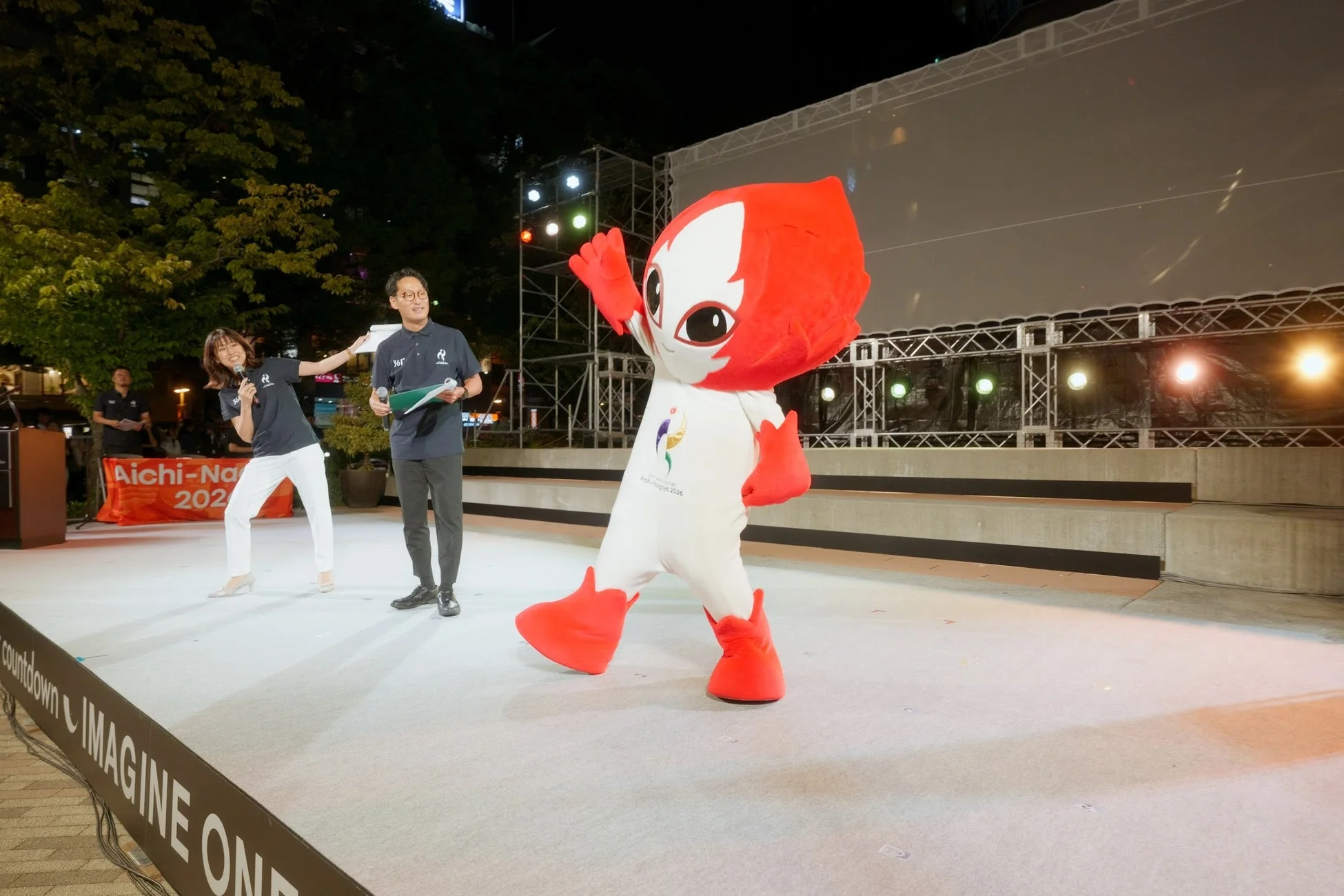 愛知・名古屋2026のイベント会場で、マスコットキャラクターと司会者がステージに立っている夜の様子