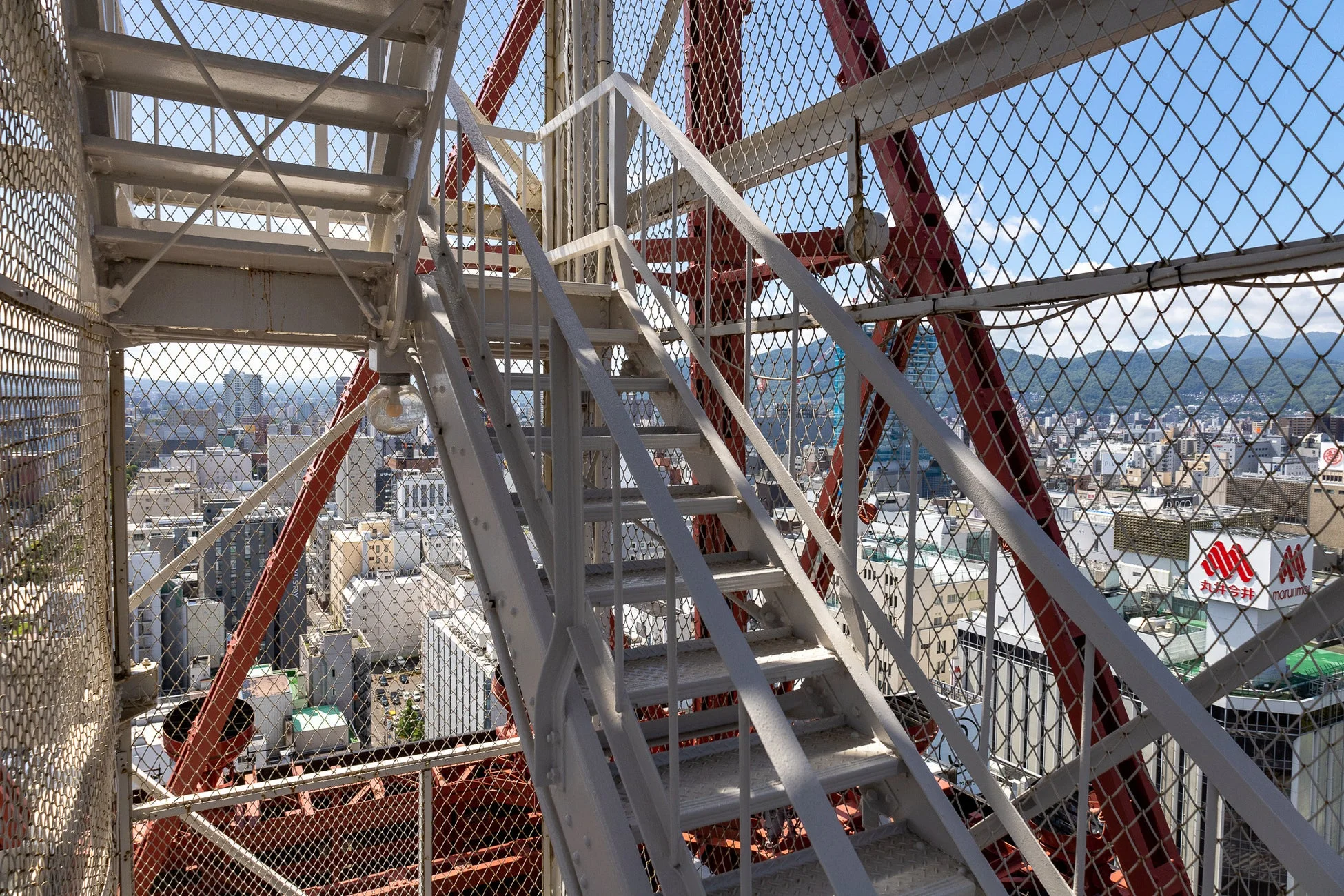 高層建築物から都市の街並みを望む風景。手前には白い金属製の階段と金網があり、奥にはビル群、遠景には山々が広がる。