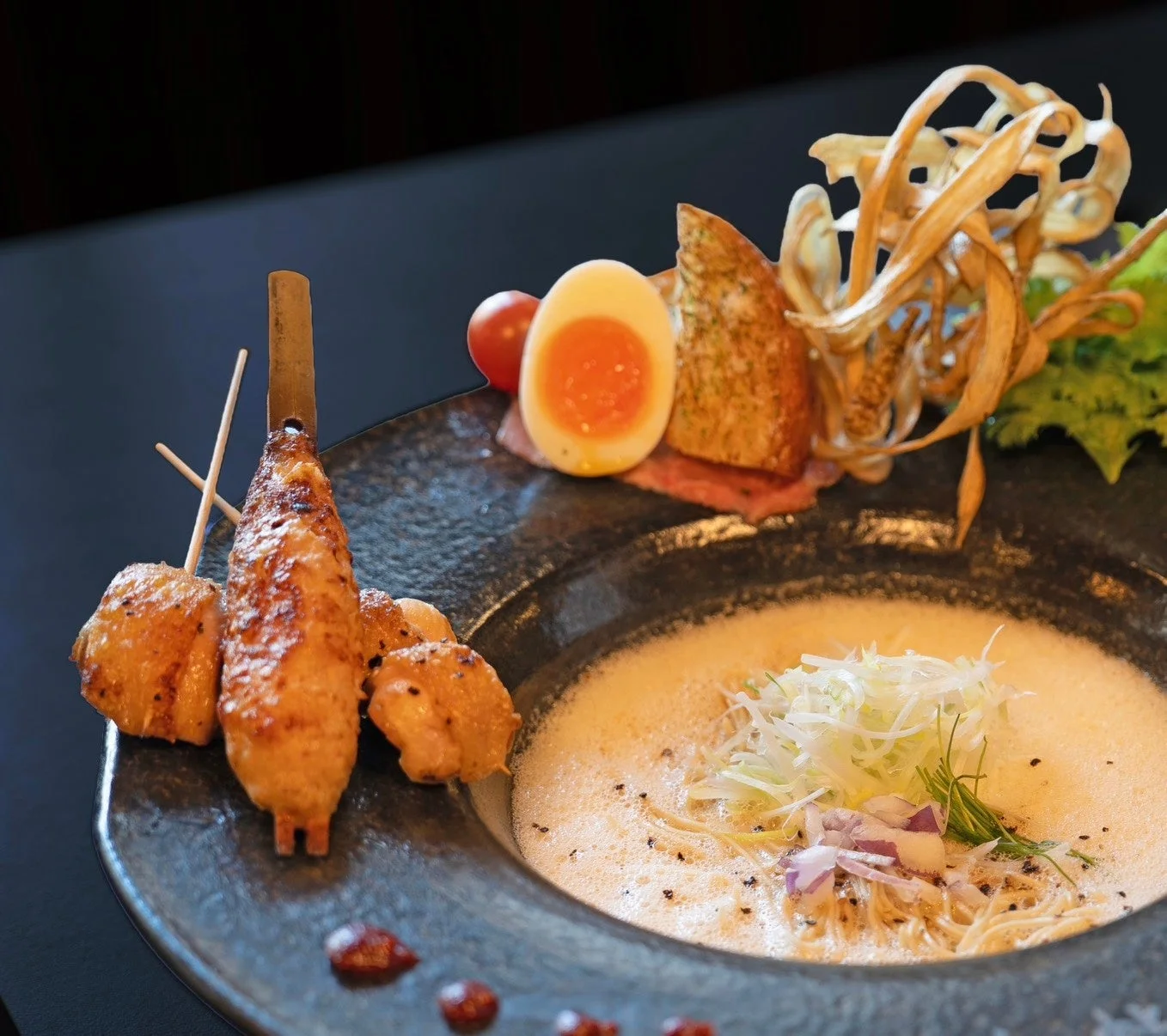 クリーミーな泡系スープのラーメンがメインの豪華な料理。鶏つくね串、半熟卵、ミニトマト、揚げ野菜などが添えられ、洗練された盛り付けが特徴です。