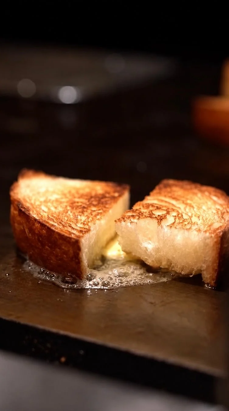厚切りのパンが鉄板の上でバターと共に焼かれ、香ばしい焼き色がついています。溶けたバターが泡立ち、食欲をそそる調理風景です。