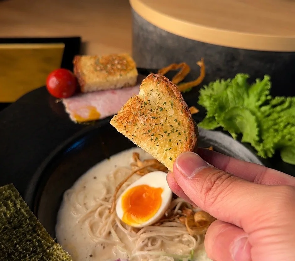 クリーミーなスープのラーメンと半熟卵がメインの皿に、ガーリックトーストを添えた食欲をそそる一品。手でトーストを持ち、まさに食べ始めようとしている瞬間を捉えています。