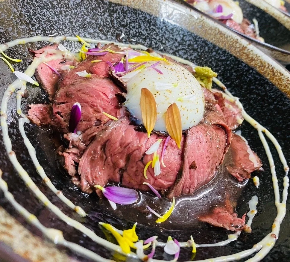 黒い器に盛り付けられた、薄切りのローストビーフに温泉卵がのった美しい一皿。